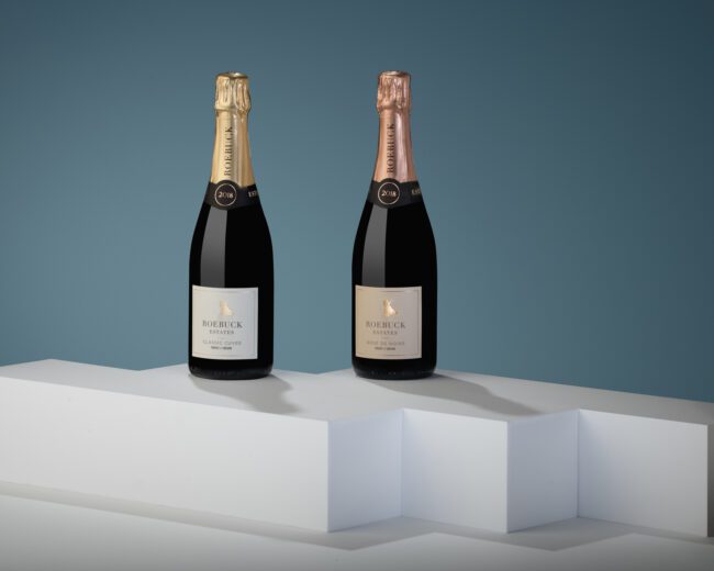 Roebuck celebration duo - Two bottles of Roebuck sparkling wines, sitting on a white platform against a dark blue background.